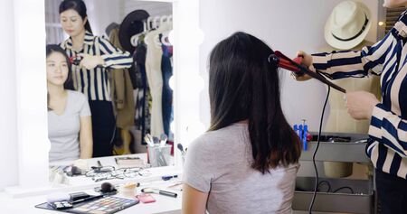 skilled young asian chinese stylist wrap curling hair in dressing room. girl actress getting ready before performance on backstage doing hairstyle. woman entertainer sit looking in mirror reflectionの写真素材