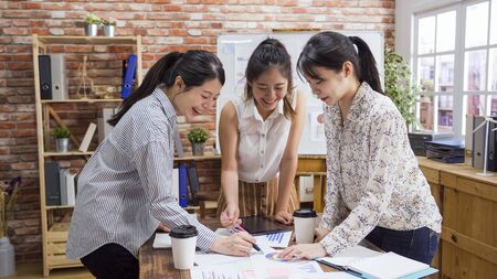 Smiling millennial people standing at shared office desk look at paperwork discuss issues together. happy company interns collaborate engaged in team brainstorming. group of women teamwork concept.の写真素材