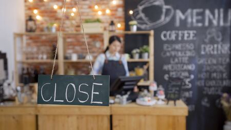 close sign black board plate hanging on window door in front. blur view bokeh of elegant asian waitress in denim apron working in background closing shop finish job in a day using pos machine counterの写真素材