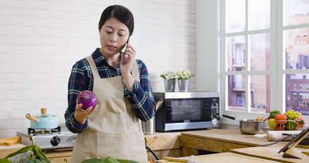 During active conversation. young beautiful asian lady holding fresh vegetables while talking on mobile phone in modern kitchen counter. Woman cooking salad and chatting on smart phone at wood tableの写真素材