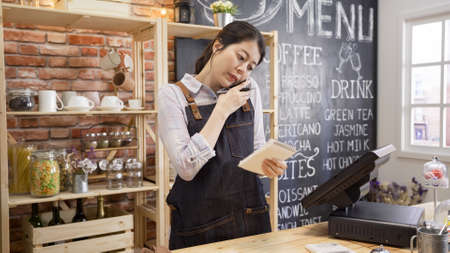 small business communication people and service concept. waitress in denim apron calling on smartphone at bar counter in coffee shop. coffeehouse staff holding note talk with customer taking orderの写真素材