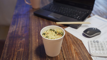 Cup instant noodle on working table in dark home kitchen. document plan papers by computer laptop...