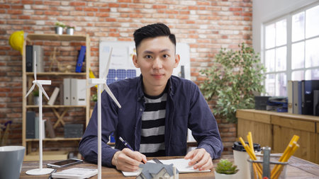 Confident asian business man looking at camera smiling with models of windmills turbines on wood...