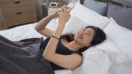 portrait chinese girl just walking up in bed is typing and sending message with smartphone looking...