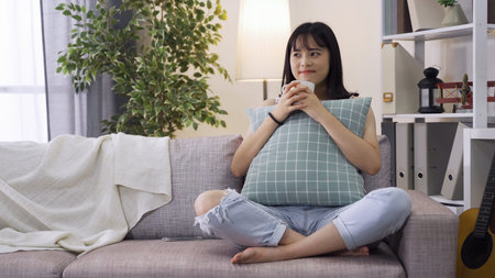 asian lady having coffee in a relaxing mood on living room couch. Young lady in love holding cushion...