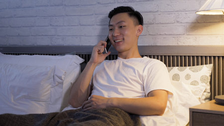 portrait smiling asian young man is speaking on phone at bedtime. chinese male sitting on bed is...