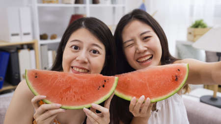 Summer indoors lifestyle concept. young girls having fun taking selfie with watermelon. beautiful female best friends smiling make self photo with red fresh fruit at home relax with funny face cameraの写真素材