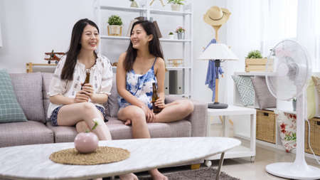 two happy smiling woman sitting near ventilator on sofa having fun gossip. girl friends chatting drinking bottle beer on couch in living room at home with electric fan open keep air cool i house.の写真素材