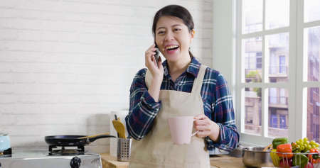 beautiful asian japanese mom in apron hold cup of morning tea and talking on mobile phone while standing in kitchen. joy lady laughing while having conversation on cellphone. smiling female at homeの写真素材