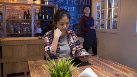 asian female customer choosing wine from alcohol list on mobile touch pad. small business ownership entrepreneur concept. girl waitress walking to client and take order while woman read tablet menuの写真素材