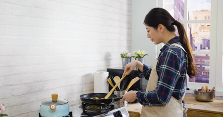 side view portrait of young asian korean woman in apron frying vegetables into pan in kitchen....