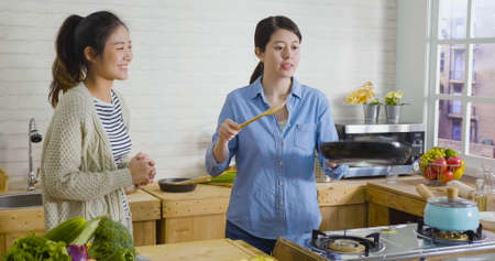 two happy girl friends cooking and having fun in cozy wooden style kitchen. women preparing food together and laughing. charming lady flipping egg in pan while roommate smiling watching performanceの写真素材