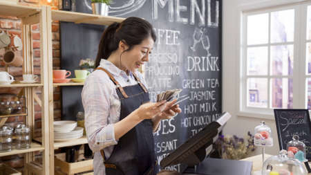 female businesswoman owner of cafe bar counting finances using calculator in small coffeehouse. smiling girl coffeehouse staff joyful with earning money in counter. waitress doing close shop routineの写真素材