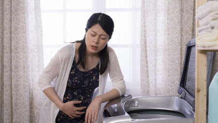 portrait female experiencing sudden labor contraction pain as she is gathering clean clothes. pregnant woman leaning against washing machine, holding belly, suffering great discomfort.の写真素材