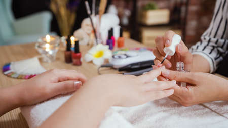 close up view asian chinese female manicurist applying transparent base coat to customer lady nail. woman beautician holding brush painting nail varnish polish on client girl hands in beauty salon.の写真素材