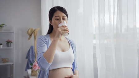 Young pregnant woman with glass of milk standing in bright room by window. elegant maternity lady drinking fresh healthy beverage in house. happy future motherhood thirsty enjoy leisure time at homeの写真素材
