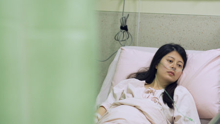 view behind hospital curtain asian woman with brain severely damaged is lying on bed. camera movement portrait of awake patient in ward with lifeless facial expressionの写真素材