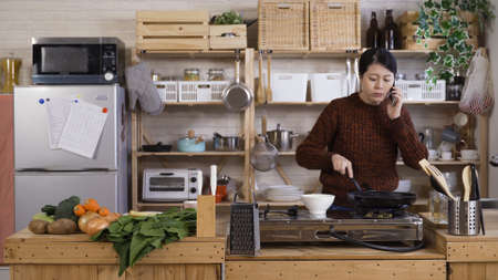 occupied asian career woman is dealing with work on mobile while cooking at home. busy morning chinese female manager holding phone is assigning work, making breakfast, and trying to find time to eatの写真素材