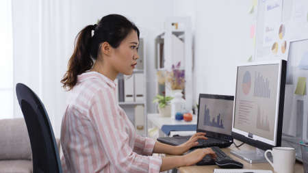 taiwanese female manager analyzing financial performance of company on computer and marking down briefly on paper. portrait of asian lady working from home studio.の写真素材