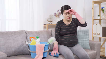 asian pregnant woman sitting on sofa hands in gloves wiping sweat off, doing too much housework at home. overworked during pregnancy concept. real moments.の写真素材