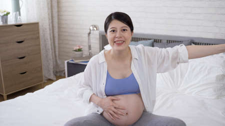 health and fitness female vlogger is touching her swollen belly and stretching her arm. pregnant woman wearing sportswear bra is looking at camera and talking to her audienceの写真素材