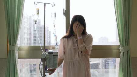 portrait of hospitalized female cancer patient back on window holding iv drip stand and weeping in despair.の写真素材
