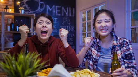 two happy young asian chinese women friends cheering for team and smiling while spending time together in bar with bottles of beer and chips.の写真素材