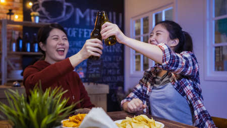 group of cheerful asian female people cheers bottles of beer and eating snacks while celebrating in night pubの写真素材