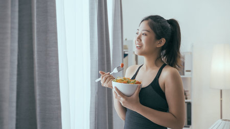 elegant asian chinese woman watching view from window and eating a bowl of healthy salad after...