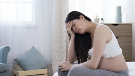 gloomy korean mother-to-be with naked abdomen sitting on bed and feeling depressed with one hand covering forehead.の写真素材