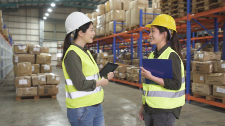 Two warehouse asian female managers in protective workwear organizing distribution standing in warehouse storage area.の写真素材