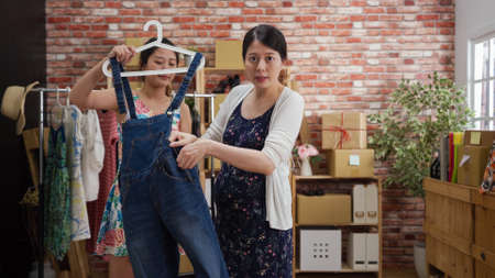 confident pregnant woman seller face camera on live streaming talking and speaking while young girl assistant helping taking denim overallsの写真素材