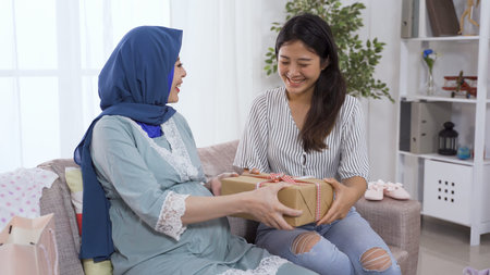Islamic expectant mother receiving gift from her female friend while talking happily on living room sofa.の写真素材