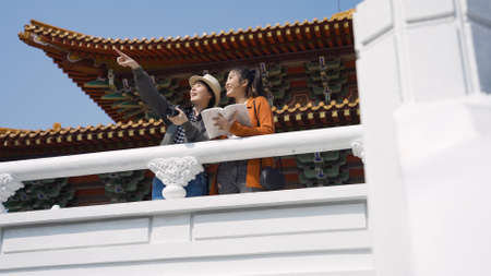 low angle shot women visitors leaning against white handrail outside a temple are pointing afar and holding a guidebook.の写真素材