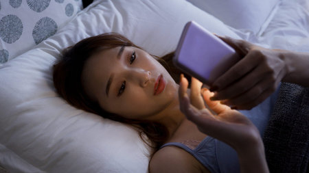 Chinese woman lying on white bedding at home is phubbing by sliding and looking at the phone screen...