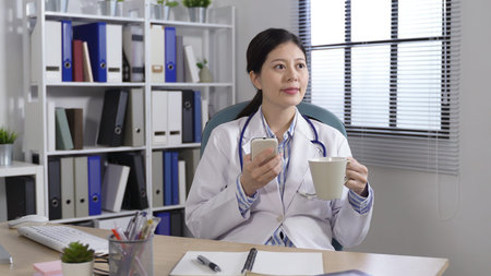happy physician relaxing on the office chair with a cup of coffee is glancing at the distance while...