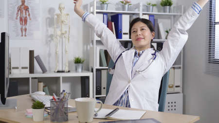 cheerful female medical worker is resting in her office chair with hands behind head while lifting her arms and shouting for joy after completing workの写真素材