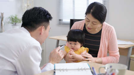 kind male physician is interacting with the adorable little baby girl by using a pen as she is sitting in her momâs arms during a consultation at clinic.の写真素材