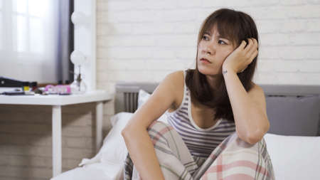 portrait sad and depressed young asian korean girl sitting and holding head in comfortable white bed in the morning in low spirit.の写真素材