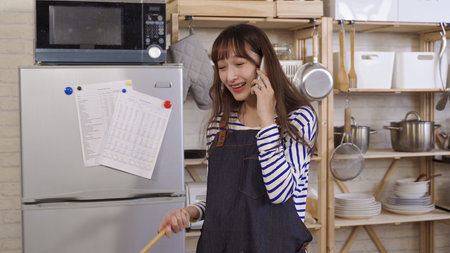 young asian female cooking in kitchen having pleasant phone talk. taiwanese lady wearing apron walking away from stove with wooden spoon in hand, burst out laughing. genuine lifestyleの写真素材
