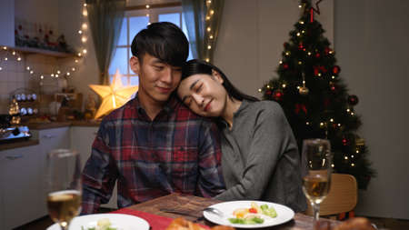 Beautiful passionate couple having romantic dinner at home celebrating christmas at night. young woman and husband hugging together by dining table with delicious xmas mealの写真素材