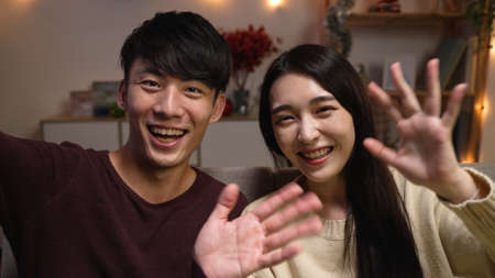 front view young asian man and woman sitting on couch at home and face camera with video call to family. cheerful lovely couple celebrating anniversary indoors and waving hands to lens and chattingの写真素材