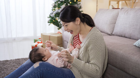 cheerful first time mom seated on the ground against the couch at home is looking at her cute baby...