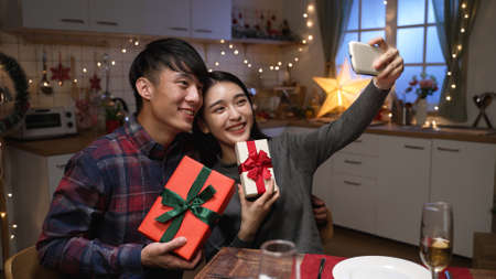Excited young couple sitting at dinner table in kitchen exchanging with gift boxes during date night on valentines day. two lovers holding present and cellphone making selfie togetherの写真素材