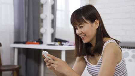 portrait asian chinese teenage girl chuckling, having fun playing mobile game at home while sitting in bed in the morningの写真素材