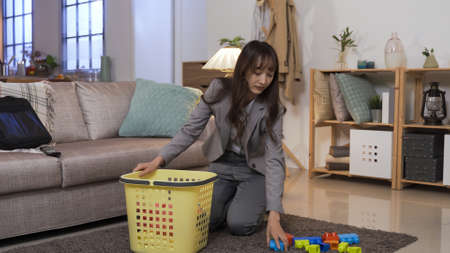 cleaning up the mess in living room. Taiwanese career mom kneeling on floor is putting toys and clothes into the basket. authentic lifestyle.の写真素材