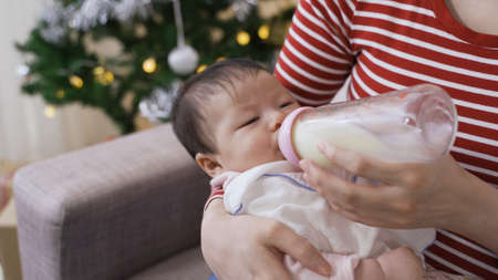 cropped shot asian innocent baby is falling asleep while lying and having bottled milk in her motherâs arms in the living room.の写真素材