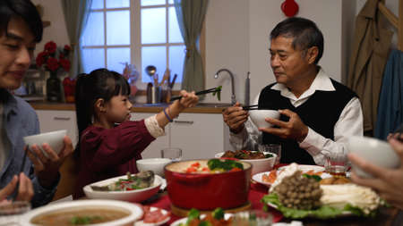 happy asian grandfather talking with hand gestures as his polite granddaughter putting food into his bowl at dinner table on chinese new yearâs eve at homeの写真素材