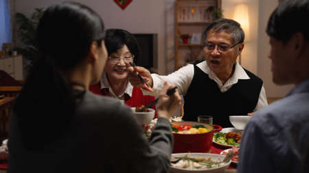 selective focus with over shoulder view smiling asian elderly couple grandfather and grandmother holding food with chopsticks and giving to their daughter in law at dinner table on eve of new yearの写真素材