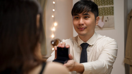 over the shoulder shot of asian man wearing suit and tie showing surprise engagement ring and...